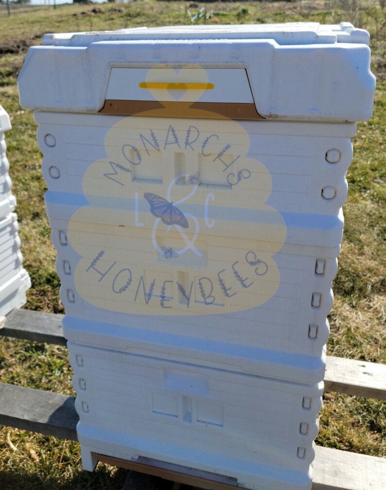 May include: White beehive with a yellow and brown top. The beehive has a label that says "Monarch & Honeybees".