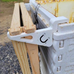 May include: White plastic beehive frame holder with a magnetic latch. The holder is attached to a white plastic beehive box. Two wooden frames are attached to the holder.