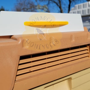 May include: A white plastic beehive entrance reducer with a yellow plastic slider. The reducer has the text "MONARCH & HONEYBEES" printed on it.
