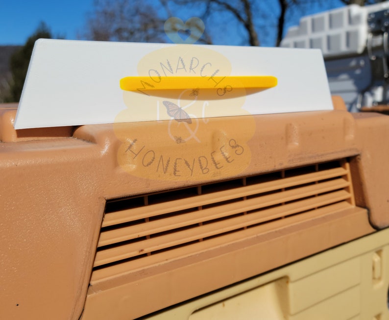 May include: A white plastic beehive entrance reducer with a yellow plastic slider. The reducer has the text "MONARCH & HONEYBEES" printed on it.