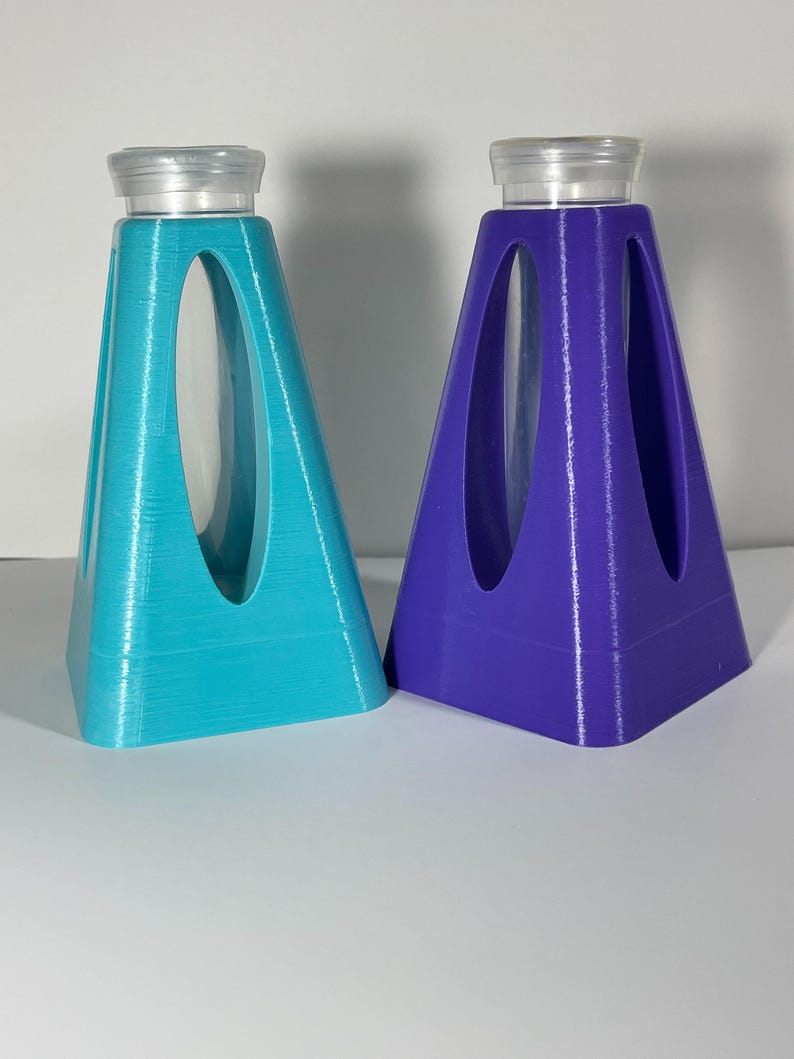 Puede incluir: Dos soportes en forma de pir&aacute;mide impresos en 3D, en color turquesa y morado, cada uno con un cilindro de vidrio transparente en el centro.