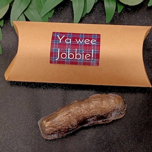 Puede incluir: Un emoji de caca marrón de chocolate envuelto en plástico transparente, con una bolsa de papel marrón con una pegatina de tartán rojo y azul que dice "Ya wee Jobbie!" en ella.