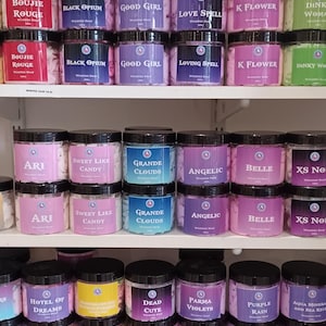 Puede incluir: Exhibición de botes de jabón batido con tapas negras y etiquetas variadas. Los botes están colocados en estantes y presentan nombres como "Cool Waters" y "Naughty Boy". Los jabones vienen en varios colores.