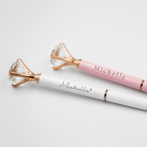 Puede incluir: Dos elegantes bol&iacute;grafos con la parte superior en forma de diamante y detalles en oro rosa. Un bol&iacute;grafo es blanco, el otro es rosa, ambos con el nombre "Michelle". Los bol&iacute;grafos son de la "Oui Collection".