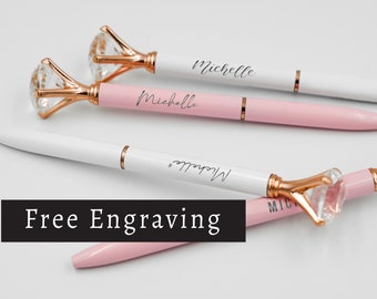 May include: Elegant pens with a diamond-shaped crystal top and rose gold accents. The pens are white and pink, with the name "Michelle" engraved on them. The image also includes the text "Free Engraving".