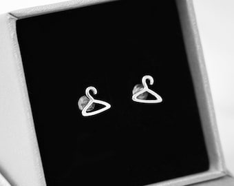 Puede incluir: Un par de pendientes de botón plateados con forma de perchas en miniatura. Los pendientes se muestran en una caja de joyería gris abierta con un interior negro. La caja tiene el texto "oui COLLECTION" impreso en la parte delantera.
