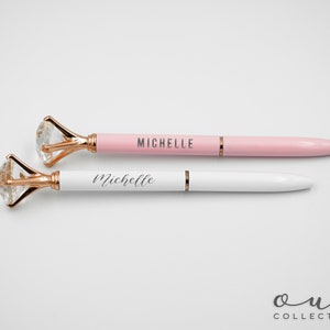Puede incluir: Dos bol&iacute;grafos con la parte superior en forma de diamante. Uno es rosa con "MICHELLE" impreso, y el otro es blanco con "Michelle" impreso. Los bol&iacute;grafos tienen detalles dorados y son de la "Oui Collection".