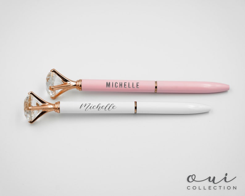 Puede incluir: Dos bol&iacute;grafos con la parte superior en forma de diamante. Uno es rosa con "MICHELLE" impreso, y el otro es blanco con "Michelle" impreso. Los bol&iacute;grafos tienen detalles dorados y son de la "Oui Collection".