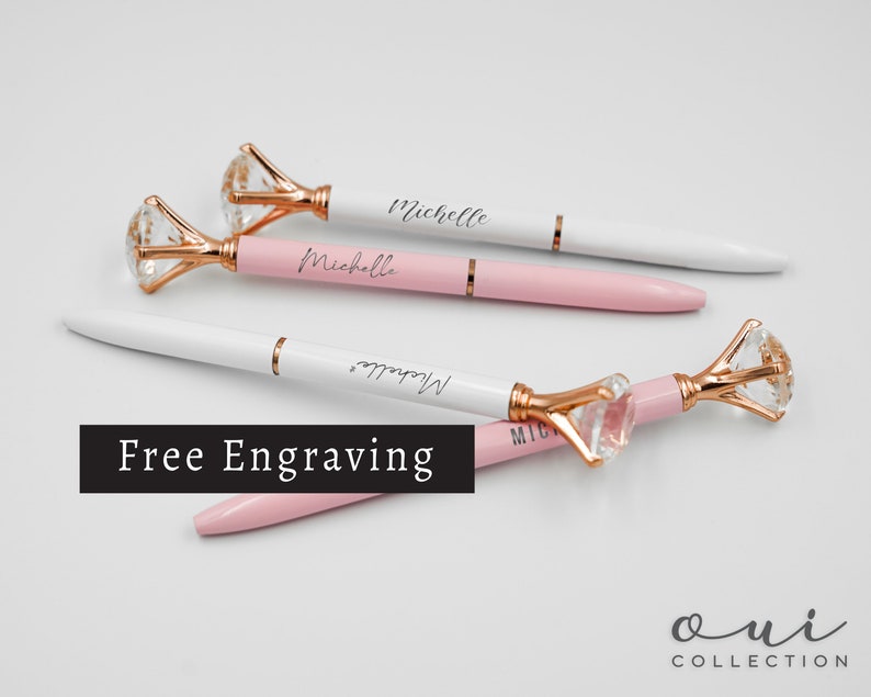 Puede incluir: Elegantes bol&iacute;grafos con una parte superior de cristal en forma de diamante y detalles en oro rosa. Los bol&iacute;grafos son blancos y rosas, con el nombre "Michelle" grabado. La imagen tambi&eacute;n incluye el texto "Free Engraving".