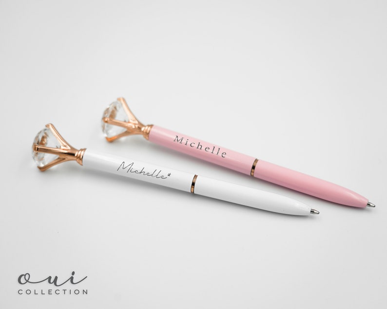 Puede incluir: Dos elegantes bol&iacute;grafos con la parte superior en forma de diamante y detalles en oro rosa. Un bol&iacute;grafo es blanco, el otro es rosa, ambos con el nombre "Michelle". Los bol&iacute;grafos son de la "Oui Collection".