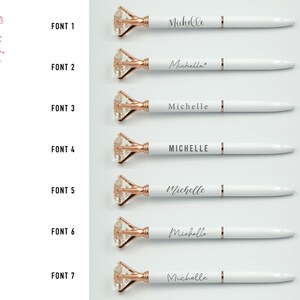 Puede incluir: Bol&iacute;grafos blancos con detalles en oro rosa y una parte superior en forma de diamante. Cada bol&iacute;grafo est&aacute; personalizado con el nombre "Michelle" en una fuente diferente. La imagen tambi&eacute;n muestra el texto "oui COLLECTION" y "FONT 1" a "FONT 7".