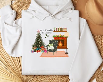 Op de afbeelding: Witte hoodie met een kerstafbeelding. Het toont een kerstboom, een open haard, een fauteuil met een kat en de tekst "ALL BOOKED for Christmas". Het ontwerp bevat ingepakte cadeaus en een boekenplank.