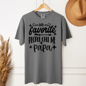 May include: Gray t-shirt with the text "My Favorite People Call Me Papa" in black script. The shirt is hanging on a wooden hanger. A brown hat is visible in the background.