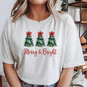 Peut inclure: T-shirt crème avec trois sapins de Noël verts ornés de nœuds rouges. L'inscription "Merry & Bright" est en rouge en dessous. Un t-shirt graphique sur le thème des fêtes.