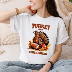 May include: A white t-shirt featuring a graphic of a brown turkey, a brown football, and a red apple surrounded by orange and yellow autumn leaves. The text "TURKEY AND TOUCHDOWNS" is printed in brown block letters above and below the illustration.
