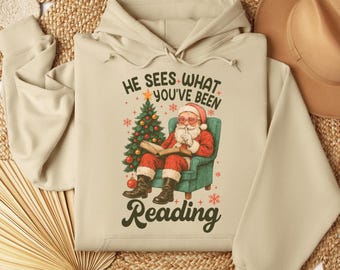 Op de afbeelding: Beige hoodie met een afbeelding van de Kerstman die een boek leest, gekleed in een rood pak en een bril. De tekst luidt "HE SEES WHAT YOU'VE BEEN READING". Een kerstboom en sneeuwvlokken zijn ook in het ontwerp verwerkt.
