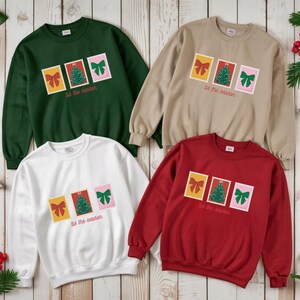 Puede incluir: Cuatro sudaderas de cuello redondo en verde, beige, blanco y rojo. Cada una presenta un diseño de tres imágenes estilo sello postal: un lazo, un árbol de Navidad y un lazo, con el texto "tis the season" debajo.