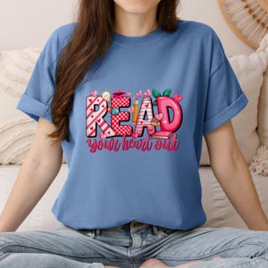 May include: A light blue t-shirt with rolled-up sleeves. The shirt features the words "READ your heart out" in a colorful graphic design. The word "READ" is in a mix of patterns and colors, with hearts and pencils.