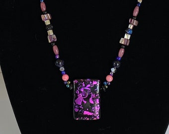 Puede incluir: Un collar de cuentas negras con un gran colgante rectangular. El colgante está hecho de vidrio morado con un patrón blanco y negro. El collar está en un maniquí negro.