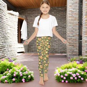 May include: A young person wearing a white t-shirt and green leggings with a pattern of yellow puppies and purple flowers. The leggings are made of a stretchy, comfortable fabric and are perfect for active wear.