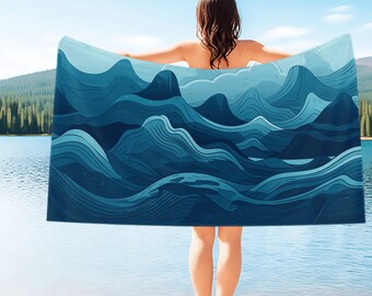May include: A person standing on a sandy beach holding a blue and white beach towel with a wave pattern. The towel is draped over the person's shoulders and the person is looking out at the water.