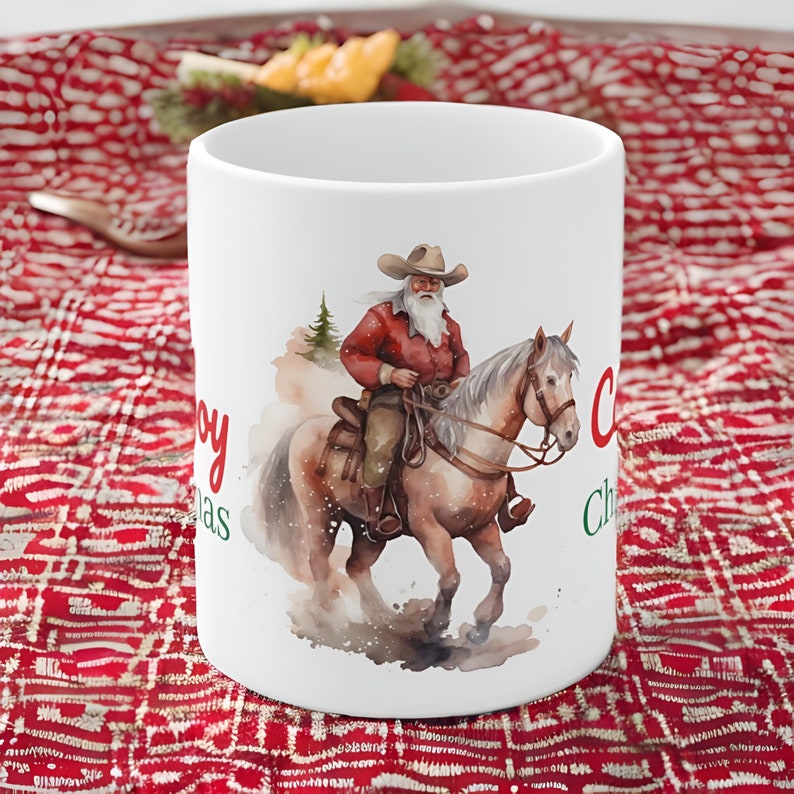 May include: White ceramic mug featuring a watercolor illustration of Santa Claus riding a horse. Santa is dressed in a red shirt and cowboy hat. The mug has the words "Merry Christmas" in red and green script.