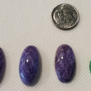 May include: Five oval-shaped cabochon gemstones, three are purple, one is green, and one is purple with a lighter shade. The gemstones are arranged in a row from left to right. A silver US dime is shown above the gemstones for size comparison.