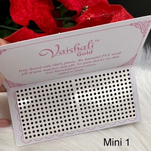 May include: A white card with pink text that reads "Vaishali™ Gold" and "Only Brand with 100% fabric. Bo harmful PVC used. pH of gum matches skin pH. No gum marks on skin Anti Microbial preservatives added to Gum". The card is folded to reveal a silver sheet with a grid of small black dots. The text "Mini 1" is printed in black at the bottom of the card.