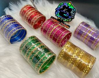 May include: An assortment of colorful bangles in red, blue, pink, green, purple, and gold. Each set is wrapped in clear plastic and features gold embellishments. The bangles are displayed on a white, fluffy surface with a decorative silver branch.