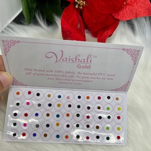 May include: A sheet of 48 small, round, adhesive, self-stick, fabric-covered, decorative, colourful, bindi dots. The bindi dots are arranged in rows and columns on a white card. The card has the text "Vaishali™ Gold" and "Only Brand with 100% fabric. Bo harmful PVC used. pH of gum matches skin pH. No gum marks on skin Anti Microbial preservatives added to Gum".