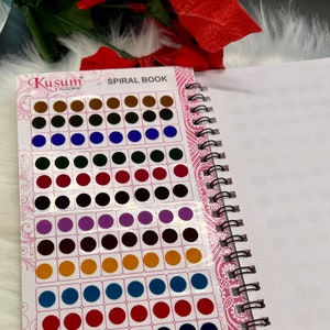 May include: A spiral-bound book of colorful bindi dots. The book is open, revealing rows of various colored dots, including brown, green, red, and blue. The cover reads "Kusum Fancy Bindi" and "Spiral Book".