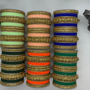 Op de afbeelding: Set van tien gouden armbanden met verschillende gekleurde stoffen inzetstukken. De armbanden zijn gestapeld in twee rijen van vijf. De kleuren van de stoffen inzetstukken zijn groen, oranje, perzik, blauw en lichtgroen.