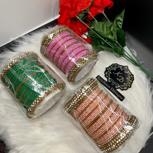 Op de afbeelding: Drie sets armbanden in de kleuren roze, groen en perzik. Elke set is versierd met gouden en heldere strass steentjes. De armbanden zijn verpakt in doorzichtig plastic en zijn gerangschikt op een wit, pluche oppervlak. Het label "Krishna Designs" is zichtbaar op de achtergrond.