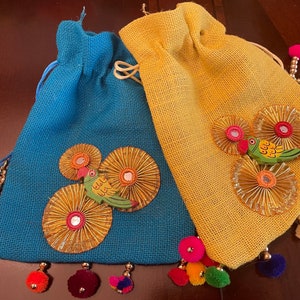 May include: Two small, decorative drawstring bags made of burlap fabric. One bag is blue with a gold and green bird design. The other bag is yellow with a gold and green bird design. Both bags have gold beads and colorful pom-poms.
