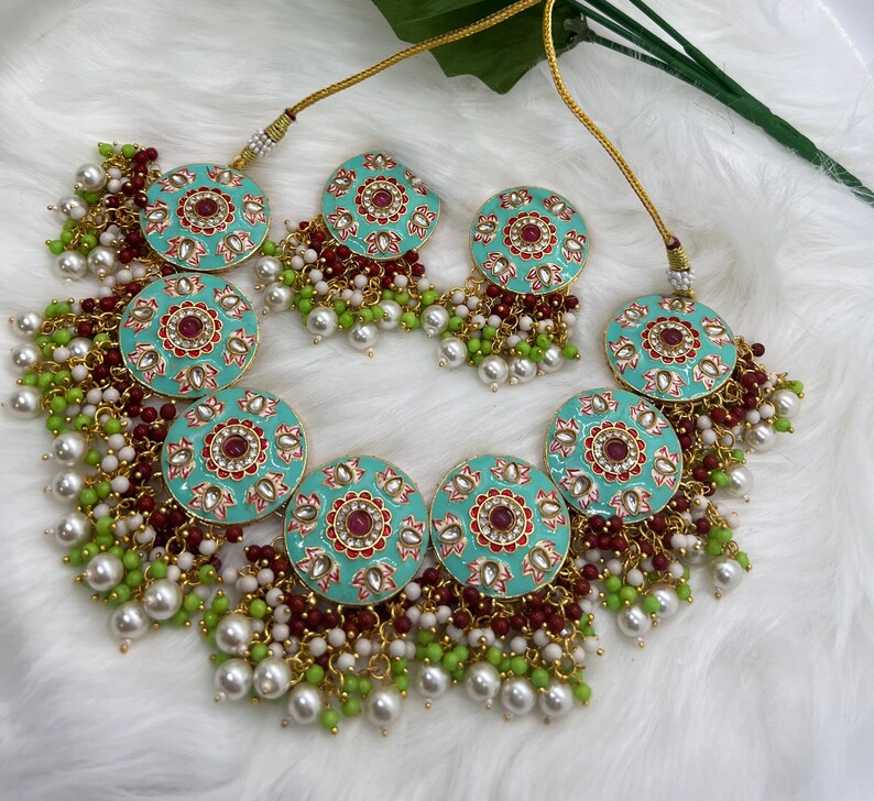 May include: A turquoise and gold necklace with red and white enamel detailing. The necklace features multiple round pendants with intricate floral designs and is adorned with white and green pearl beads.