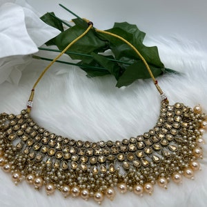Puede incluir: Un collar choker dorado con un dise&ntilde;o grande y ornamentado con piedras transparentes y facetadas. El collar est&aacute; acentuado con una franja de peque&ntilde;as perlas blancas.