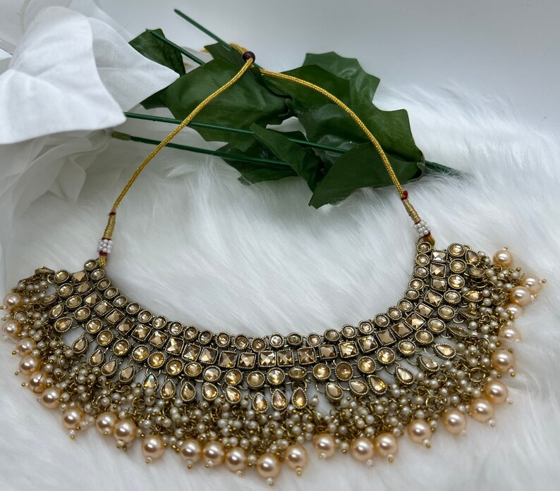 Puede incluir: Un collar choker dorado con un dise&ntilde;o grande y ornamentado con piedras transparentes y facetadas. El collar est&aacute; acentuado con una franja de peque&ntilde;as perlas blancas.