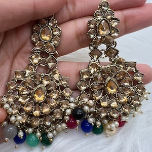 Puede incluir: Un par de pendientes de tono dorado con un dise&ntilde;o de varios niveles. Los pendientes presentan una pieza central grande, redonda, de forma floral con piedras m&aacute;s peque&ntilde;as en forma de l&aacute;grima. Los pendientes est&aacute;n adornados con una franja de perlas y cuentas de colores.