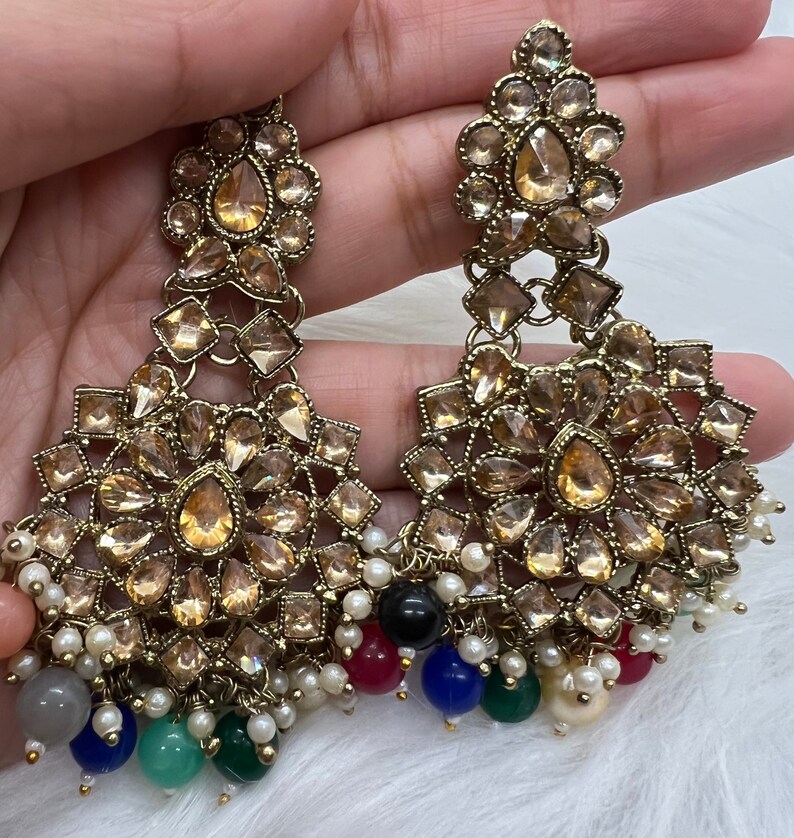 Puede incluir: Un par de pendientes de tono dorado con un dise&ntilde;o de varios niveles. Los pendientes presentan una pieza central grande, redonda, de forma floral con piedras m&aacute;s peque&ntilde;as en forma de l&aacute;grima. Los pendientes est&aacute;n adornados con una franja de perlas y cuentas de colores.