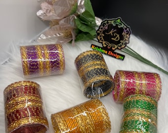 May include: Six sets of colorful bangles with gold accents. The bangles are stacked and wrapped in clear plastic. The bangles are purple, black, red, orange, and green.