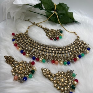 Puede incluir: Un conjunto de collar de novia indio de color dorado con cuentas de colores y detalles intrincados. El conjunto incluye un collar choker, un par de pendientes a juego y una pieza para la cabeza.