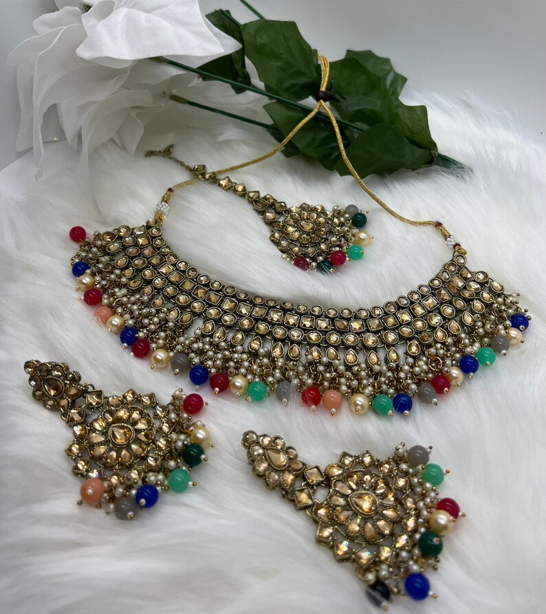 Puede incluir: Un conjunto de collar de novia indio de color dorado con cuentas de colores y detalles intrincados. El conjunto incluye un collar choker, un par de pendientes a juego y una pieza para la cabeza.