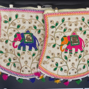 May include: Two white fabric pouches with embroidered elephants and floral designs. The pouches have colorful tassels and a zipper closure.