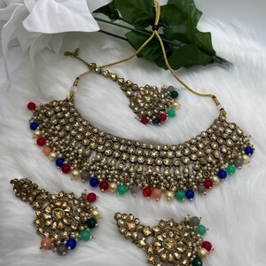 Puede incluir: Un conjunto de collar de novia indio de color dorado con cuentas multicolores y un gran colgante adornado. El conjunto incluye un collar, pendientes y un maang tikka.