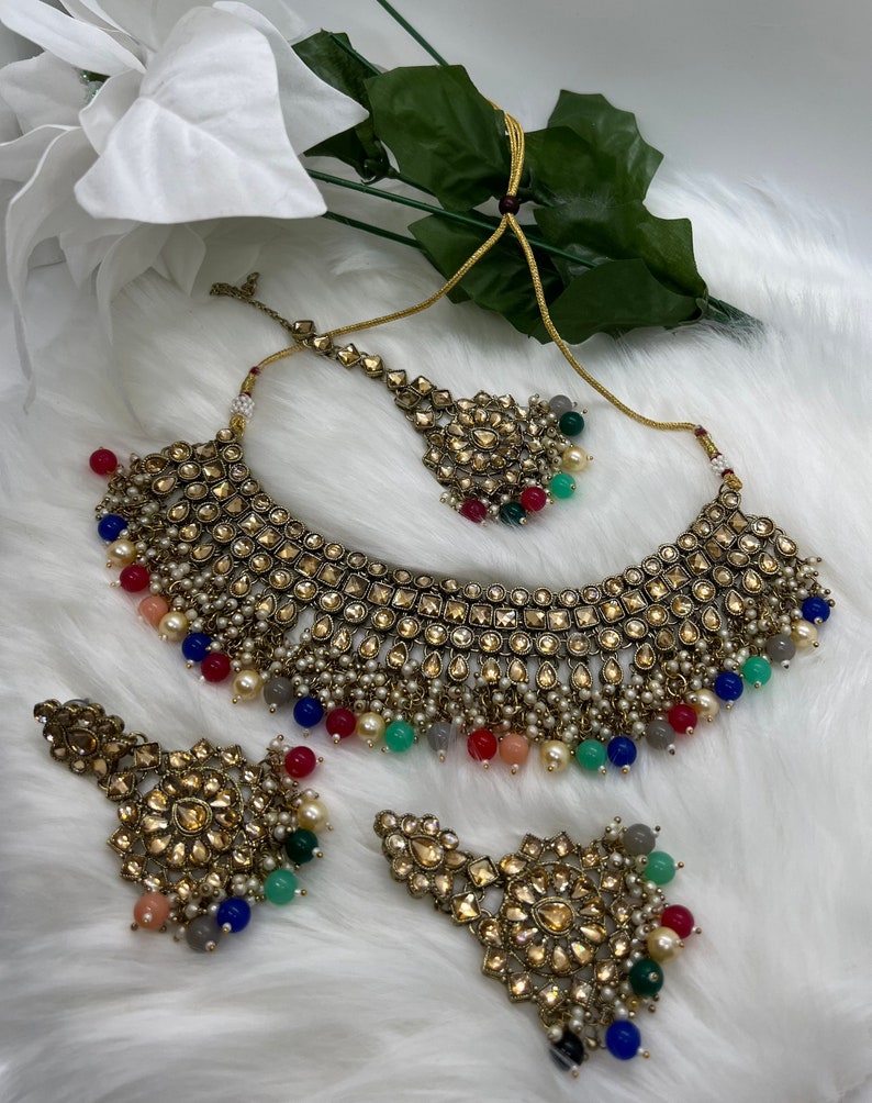 Puede incluir: Un conjunto de collar de novia indio de color dorado con cuentas multicolores y un gran colgante adornado. El conjunto incluye un collar, pendientes y un maang tikka.