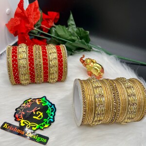 May include: Two sets of gold and red bangles with intricate designs and rhinestones. The bangles are stacked on top of each other and are displayed on a white fluffy surface. The image also includes a small gold elephant figurine.