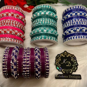 Op de afbeelding: Set van vier kleurrijke armbanden met strassversieringen. De armbanden zijn in tinten roze, groen, blauw en paars. De armbanden liggen op een wit oppervlak. De afbeelding bevat ook een logo voor Krishna Designs.