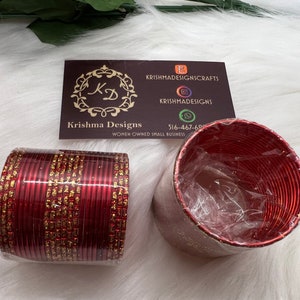 May include: A set of red and gold bangles with a glittery finish. The bangles are stacked together and are in a clear plastic bag. The image also includes a business card with the text "Krishma Designs" and "Women Owned Small Business".