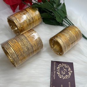 May include: Three sets of gold bangles with glitter accents. The bangles are stacked and wrapped in clear plastic. The image also includes a business card with the text "Krishna Designs" and contact information.