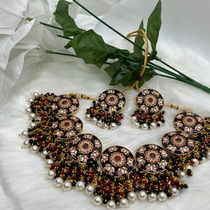May include: A black and gold necklace with red and pink floral accents. The necklace is adorned with black and white beads and pearls. The necklace is paired with matching earrings.